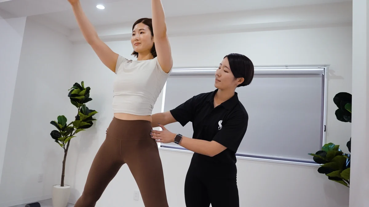 女性がリフォーマーの上で腕を伸ばし、女性トレーナーが姿勢をサポートするレッスンイメージ