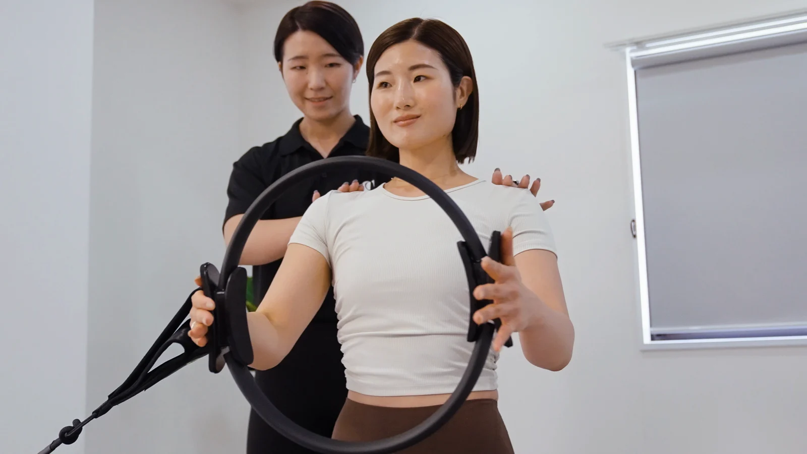 女性がピラティスリングを使い、女性トレーナーが身体の動きを確認するレッスンイメージ