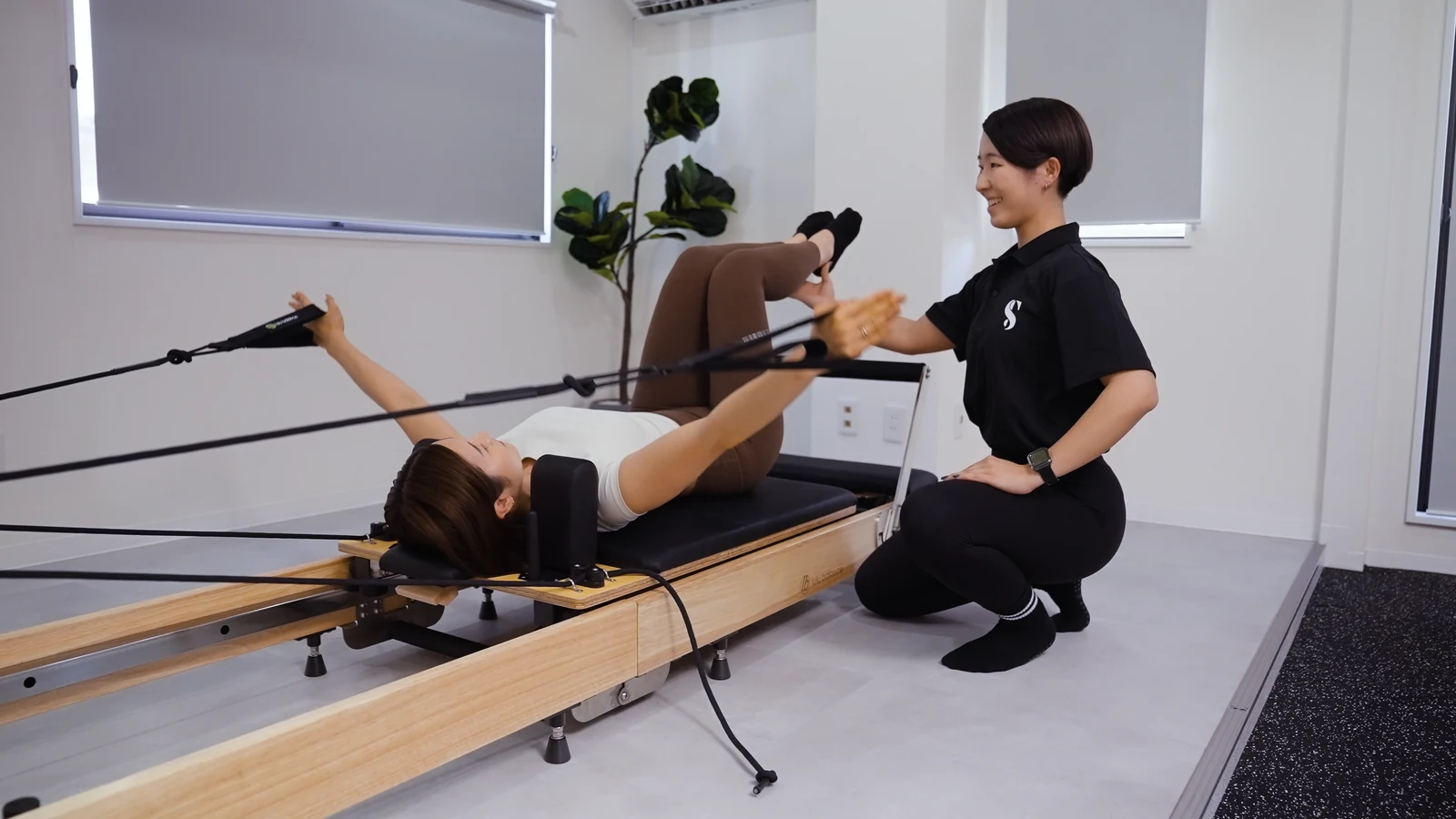 リフォーマーで腕を動かす利用者を女性トレーナーがサポートするレッスンイメージ